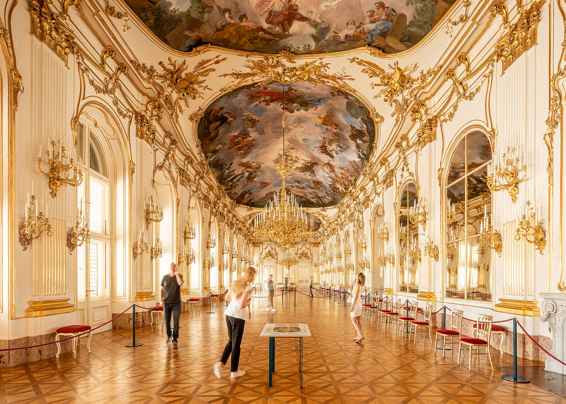 Vue de la Grande Galerie, personnes regardant les fresques du plafond, lustres dorés suspendus au plafond au centre.